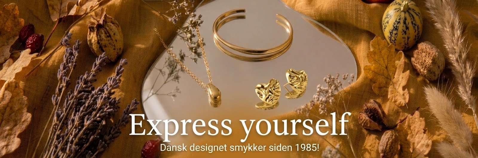 Spinning Jewelry efterårs topbanner med forgyldte smykker: Silver Dew FG halskæde, Twist armring FG og Crumble Hearts ørestikker FG præsenteret på spejl med tørrede efterårsblomster.